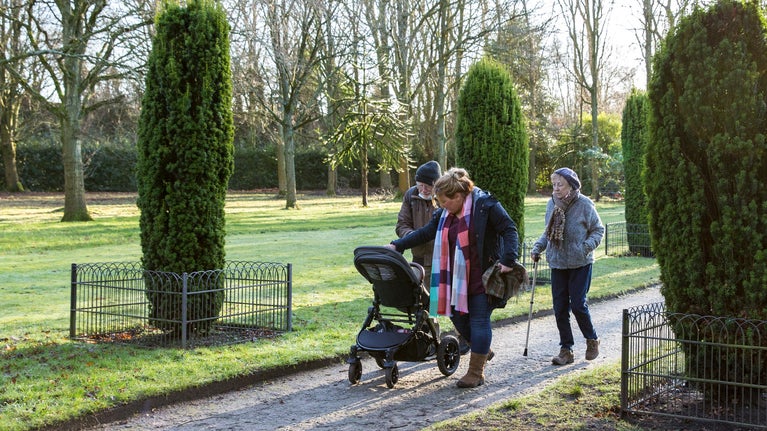 A multi-generational family of three adults and one child walk through the gardens at Speke Hall. They are dressed for winter and pushing a pram.